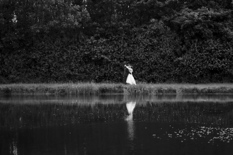 A black and white wedding photo of a couple kissing across a reservoir on their wedding day.
